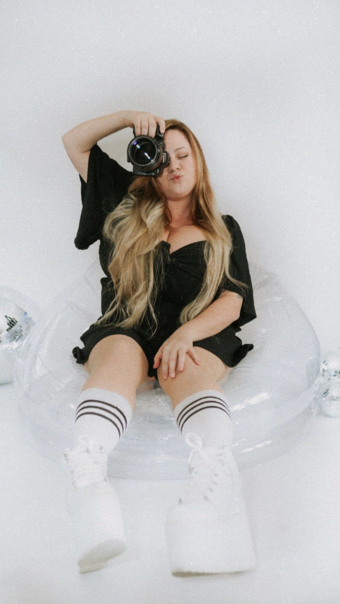 Photographer Stacia in black outfit poses with camera on clear chair with disco ball decorations. 90s nostalgic vibes spice girls shoes