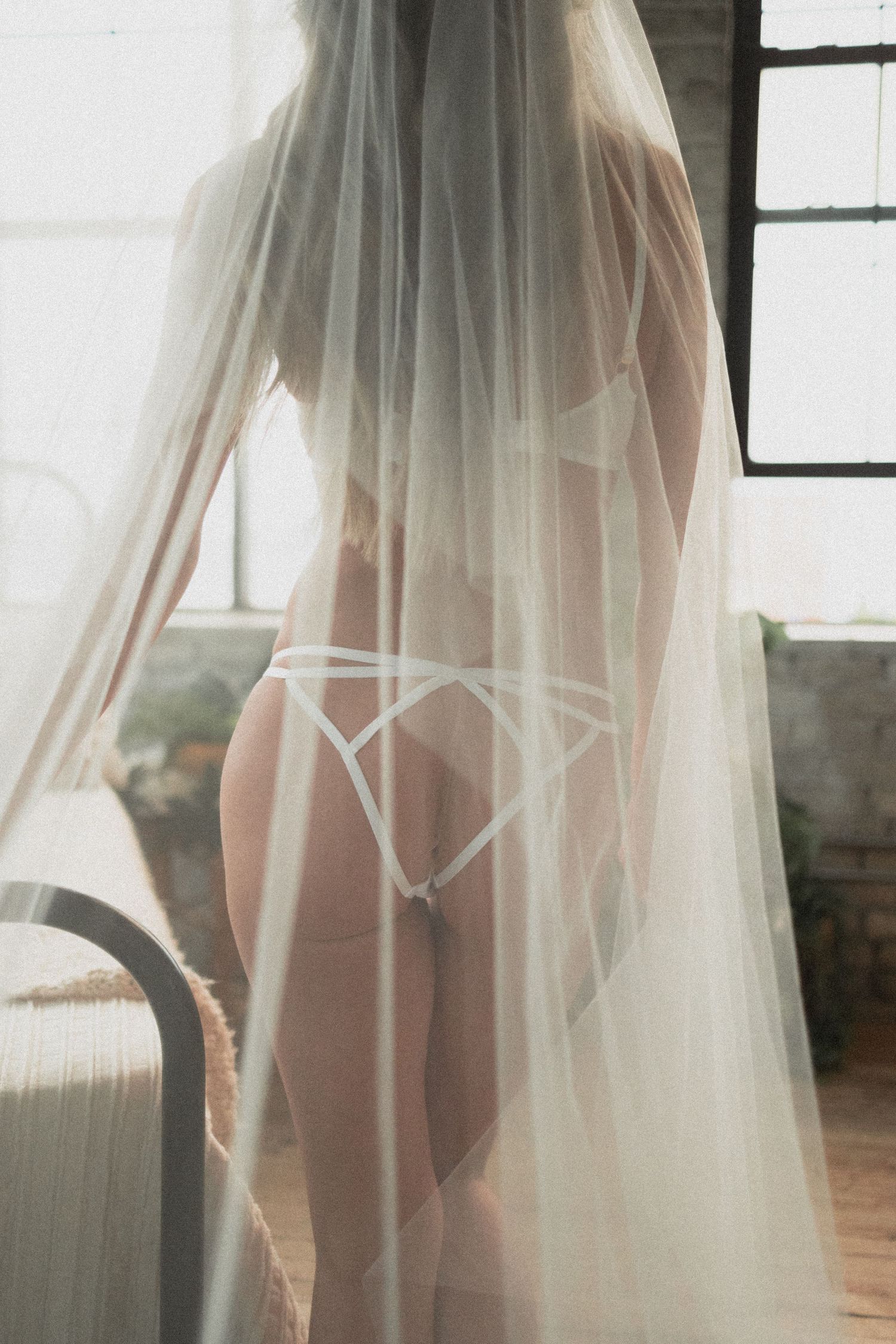 Artistic boudoir photo of person in white veil and lingerie by window.