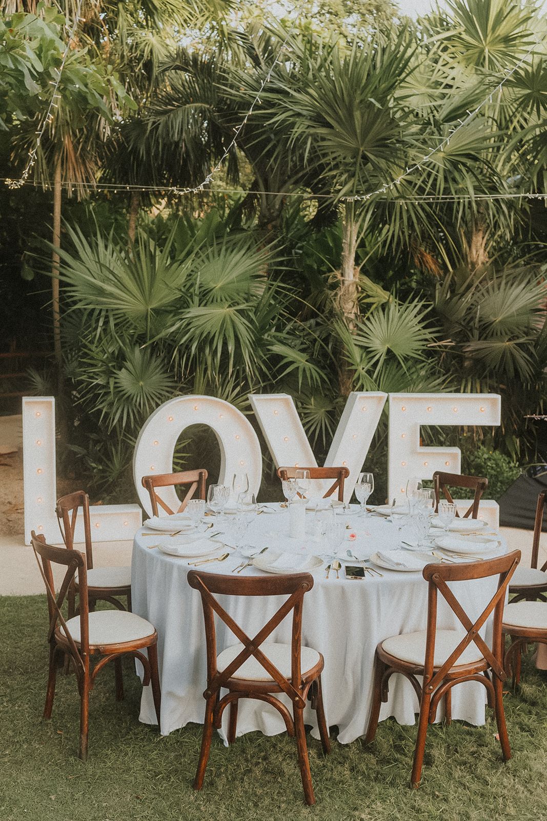Outdoor wedding reception setup with white tablecloths and wooden cross back chairs under palm trees at Secrets Akumal Mexico resort wedding