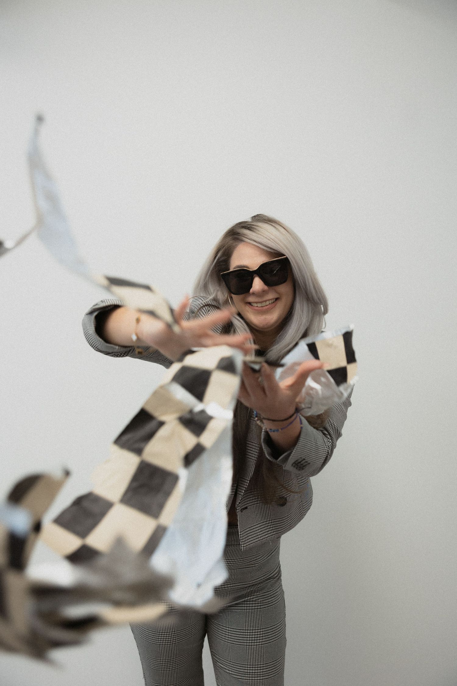 Person in gray outfit and sunglasses playfully tosses papers in the air against a light background.
