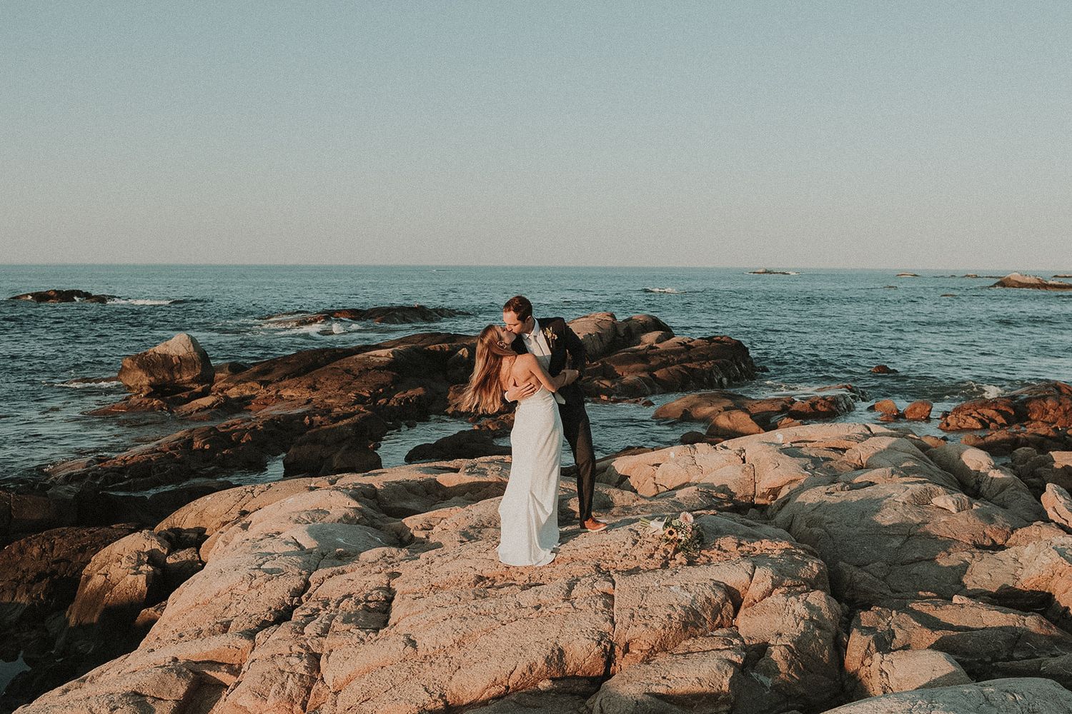 Rocky Massachusetts coastline with waves crashing against shore during golden hour, while bride and groom kiss romantically