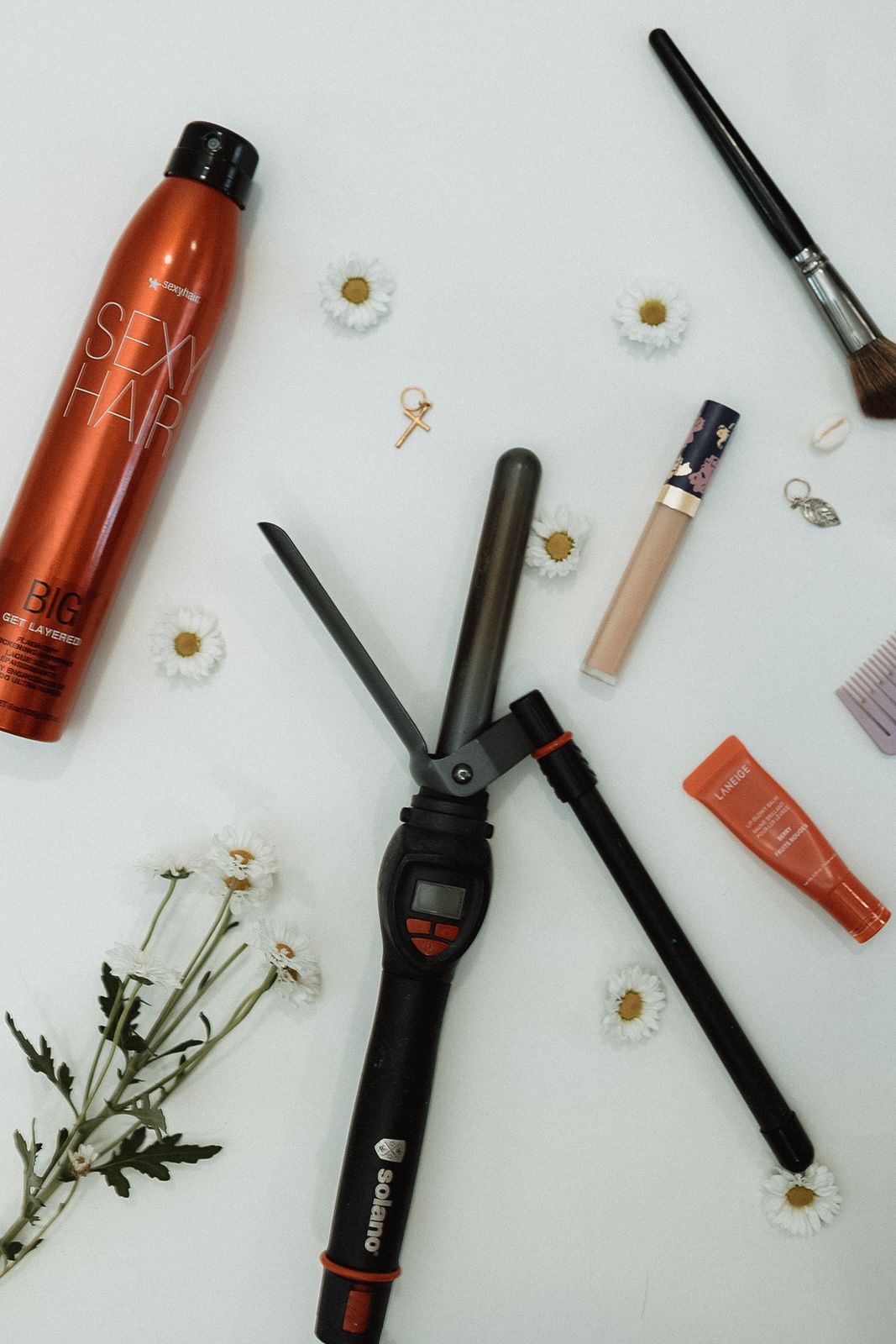 Flat lay of hair styling tools and products with daisies on white background.