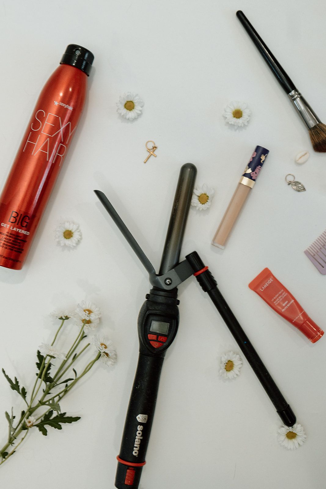 Flat lay of hair styling tools and beauty products with small white daisies scattered on white background.