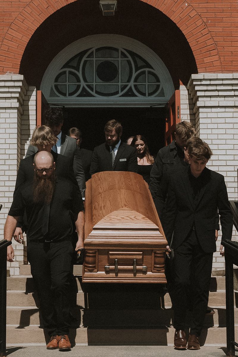 Pallbearers carrying wooden caskets outside a brick church building during a funeral service.