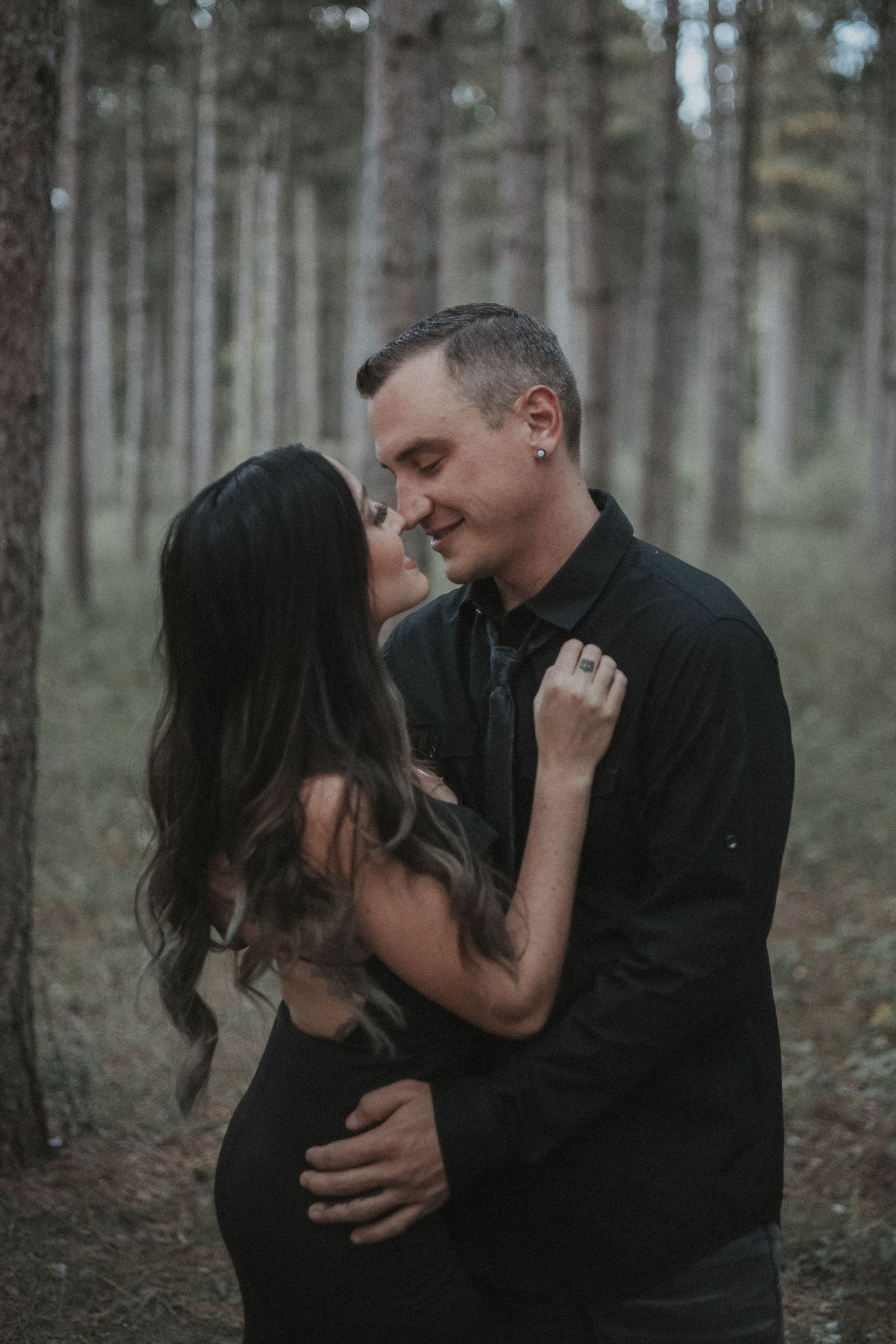 Couple in black outfits sharing intimate moments in a pine forest setting.