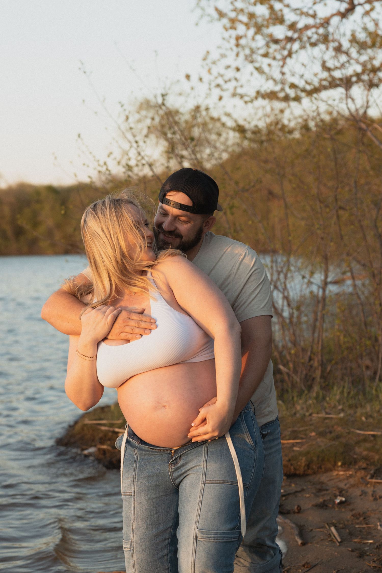 A couple embraces by a lakeside at golden hour, the woman visibly pregnant in a white crop top and jeans.