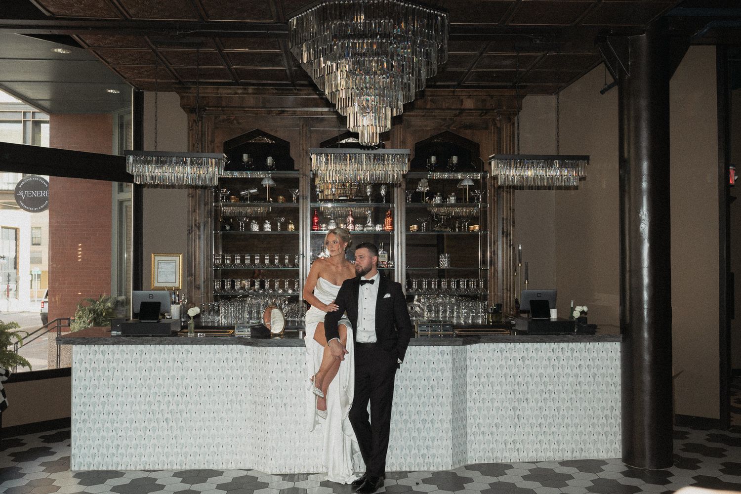 A wedding reception in a modern industrial venue with crystal chandeliers and white brick walls. Bride & Groom sitting on a bar at Le Venere