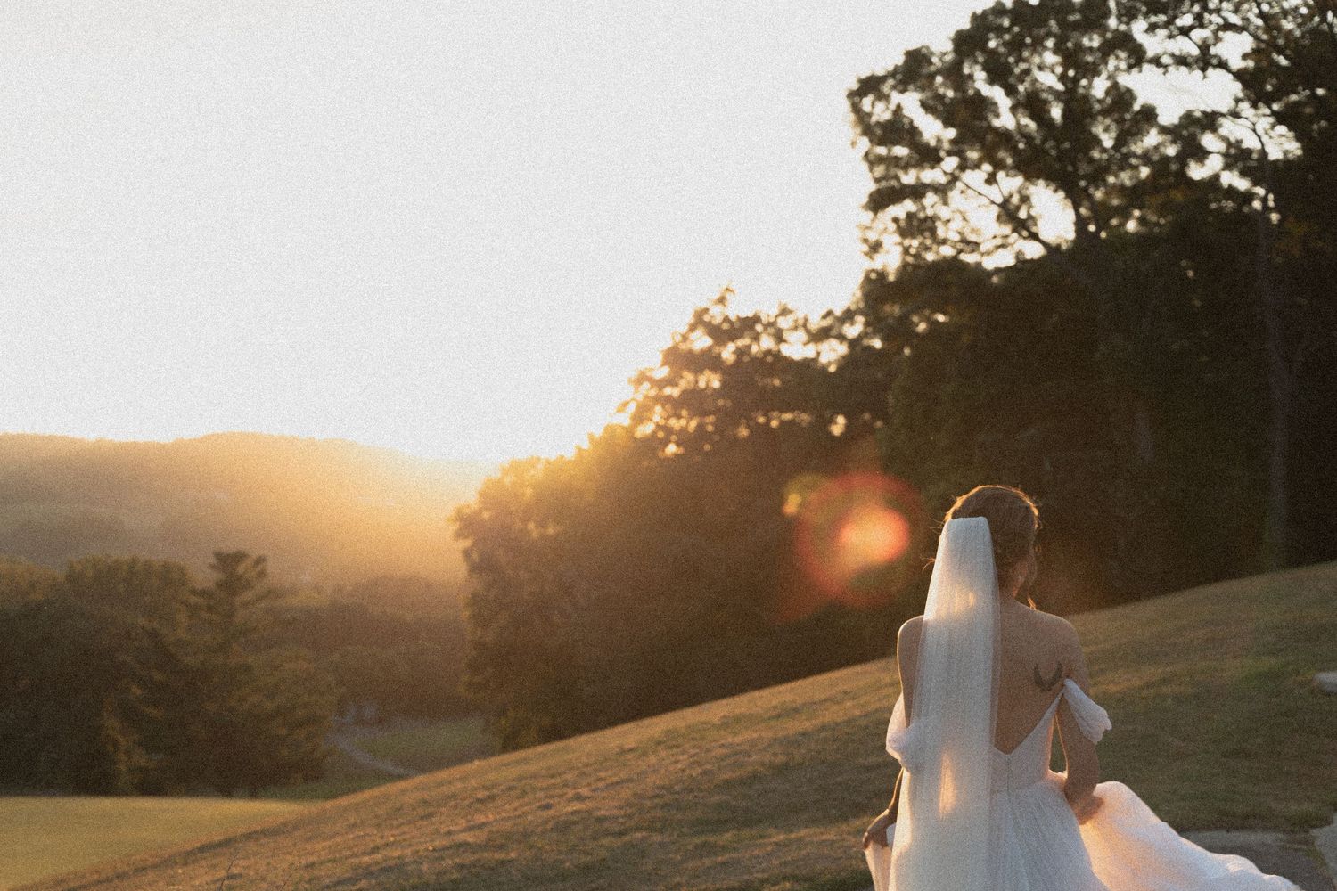 Bride in white dress stands on hillside during golden sunset with trees silhouetted in background at The SkyRoom Weddings in Redwing MN