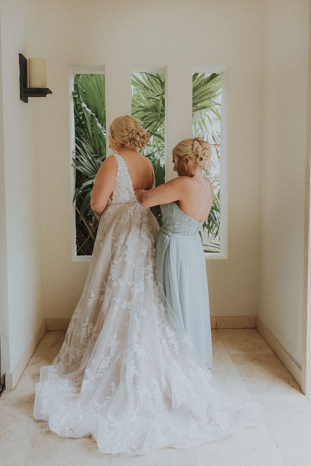 Mother of the bride helping bride into flowing white gown, standing together in an elegant hallway at Secrets Akumal Mexico resort wedding