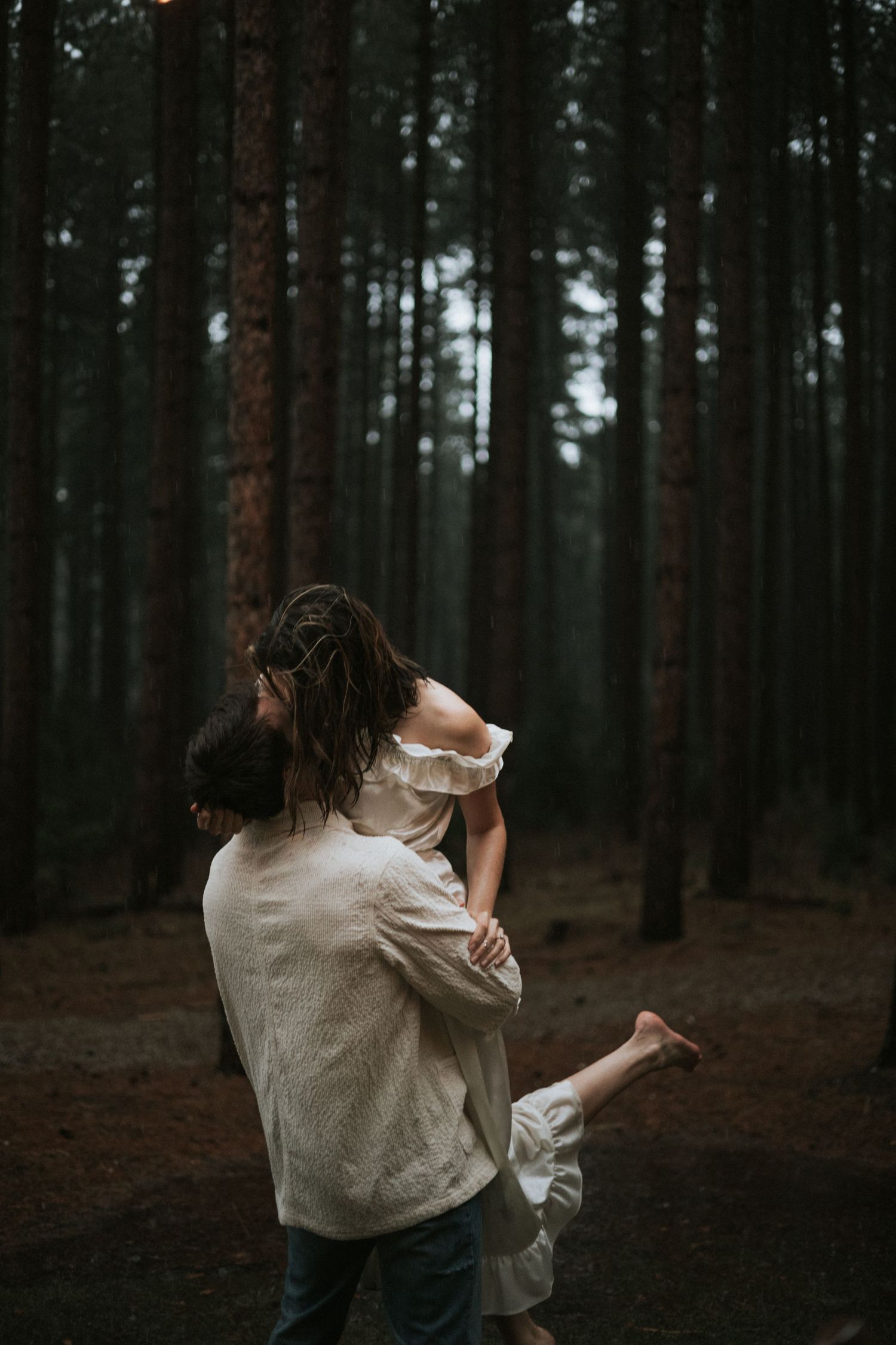 A person in a white sweater dances alone in a moody dark forest setting with tall pine trees in the background.