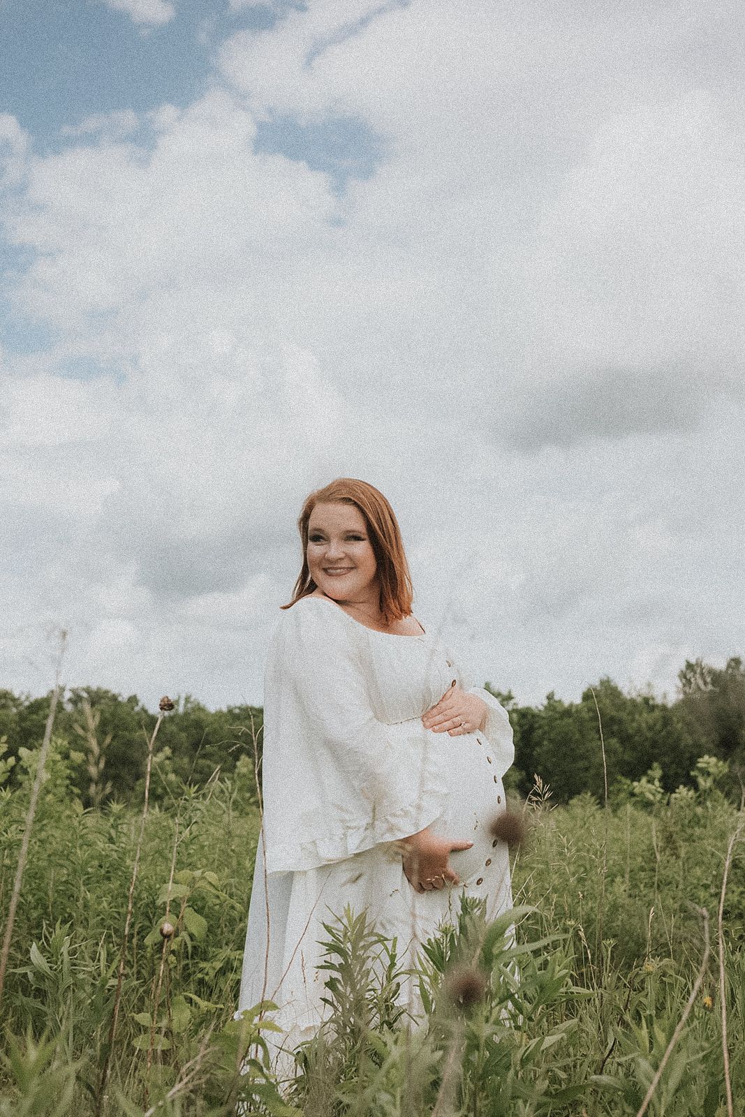 Pregnant lady in flowing white dress standing in a sunlit meadow with wildflowers and cloudy sky.