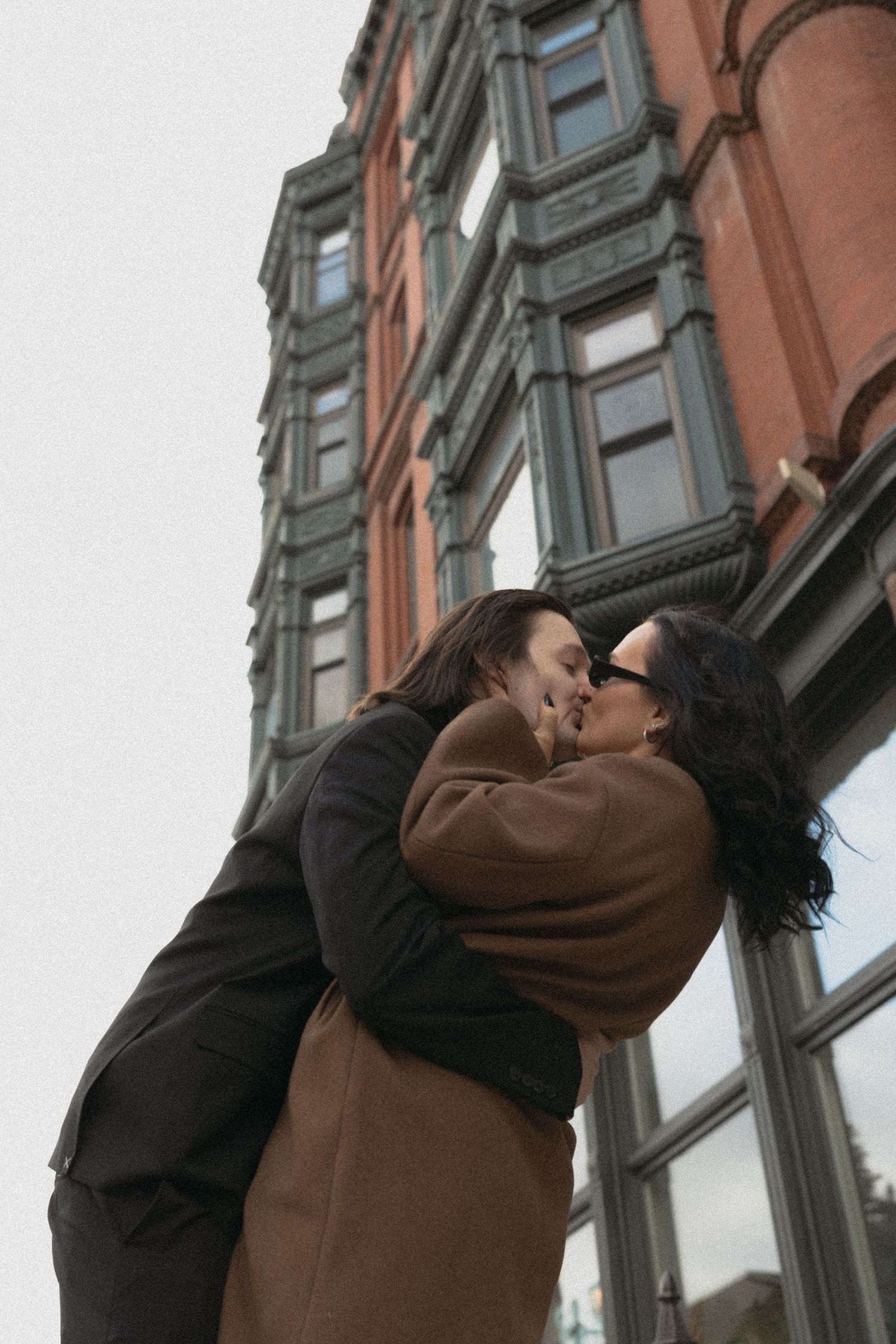 Couple sharing a romantic kiss outdoors in front of a classic red brick building on an overcast day.