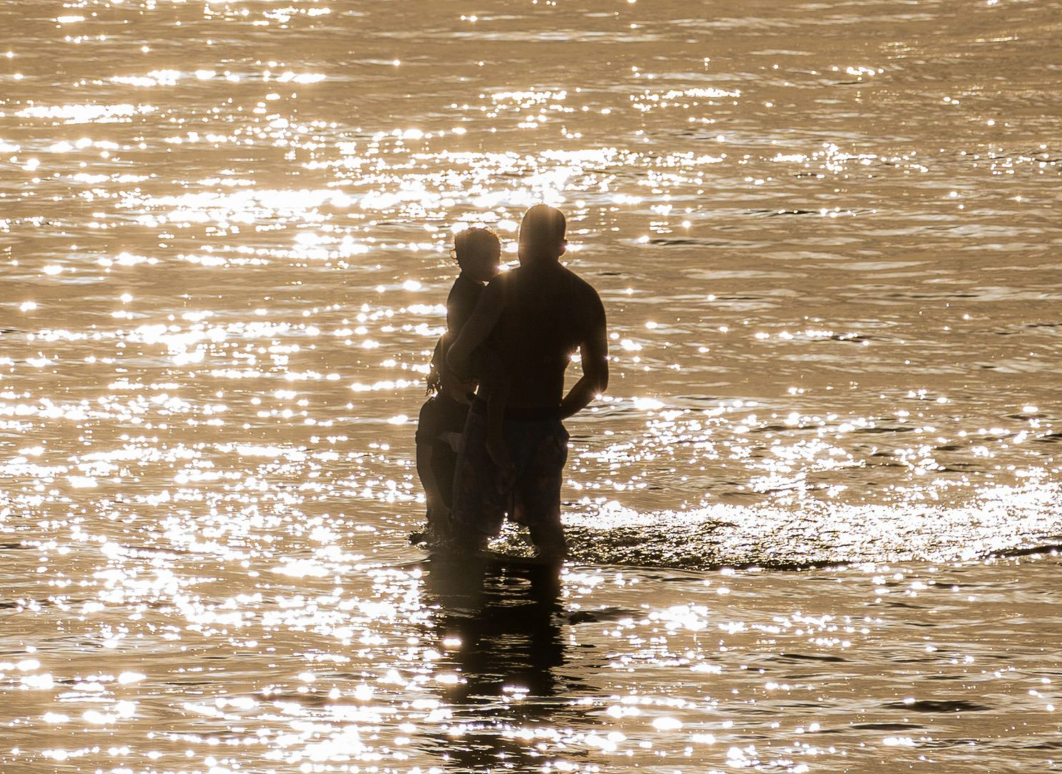 Silhouettes of people wading in sparkling golden water during sunset.