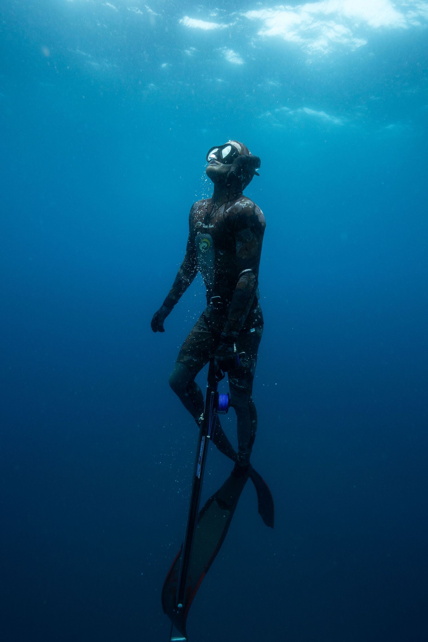 A scuba diver silhouetted against blue ocean water hovers vertically in the deep while ascending to the surface.