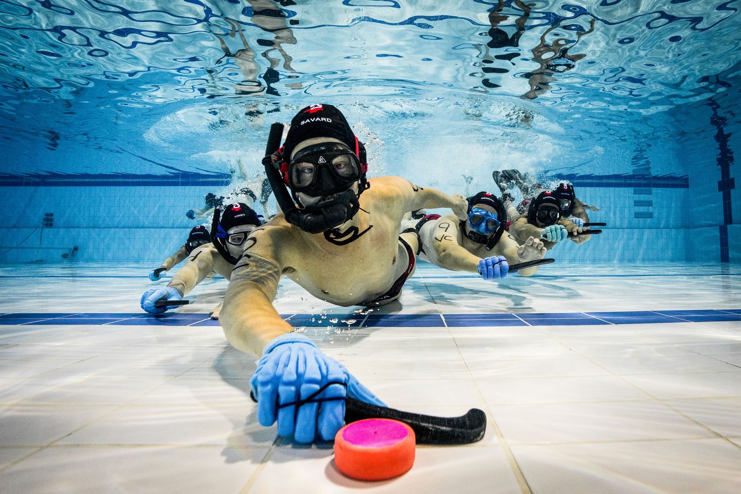 Underwater hockey players compete for the puck on the pool floor in a dynamic sports match.