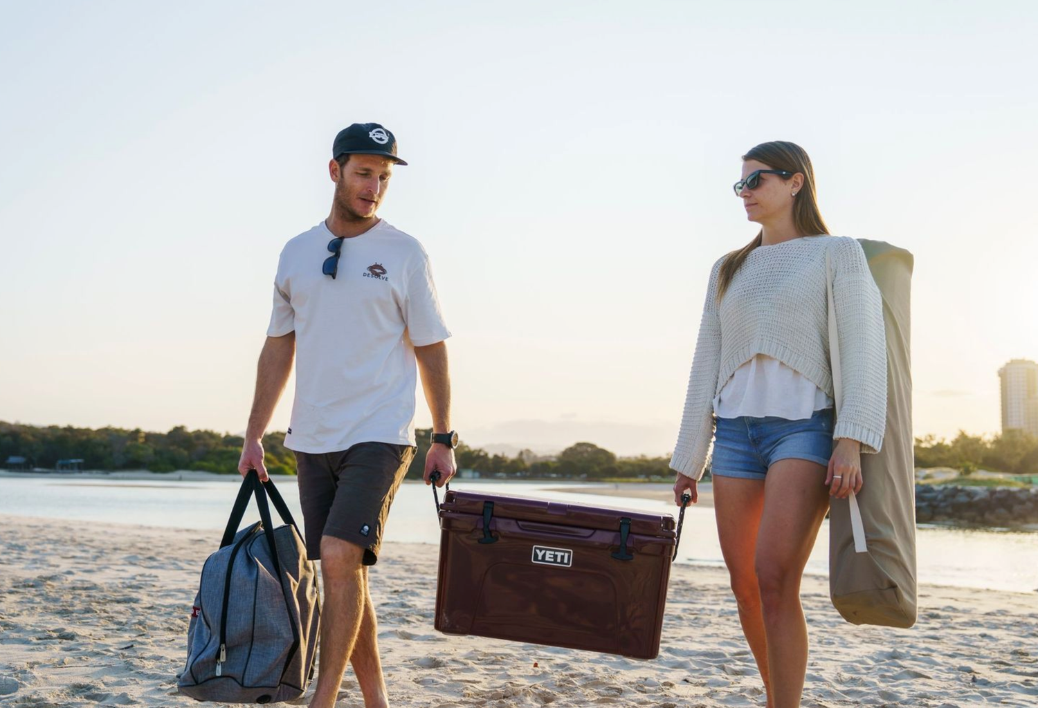 Two people carrying cooler and bags along beach at sunset.
