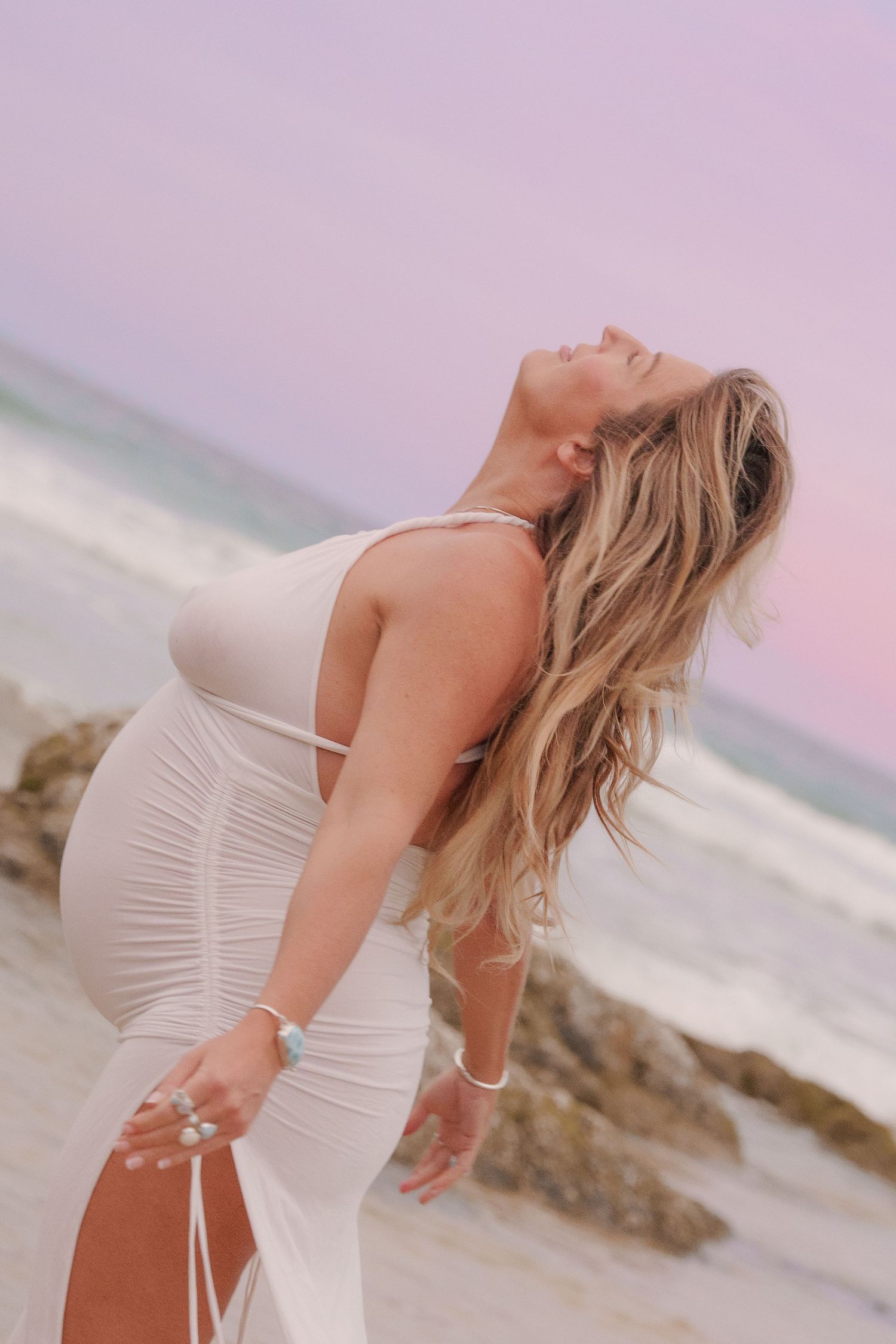 A person in a white dress poses gracefully on a beach at sunset with rocky outcrops in the background.