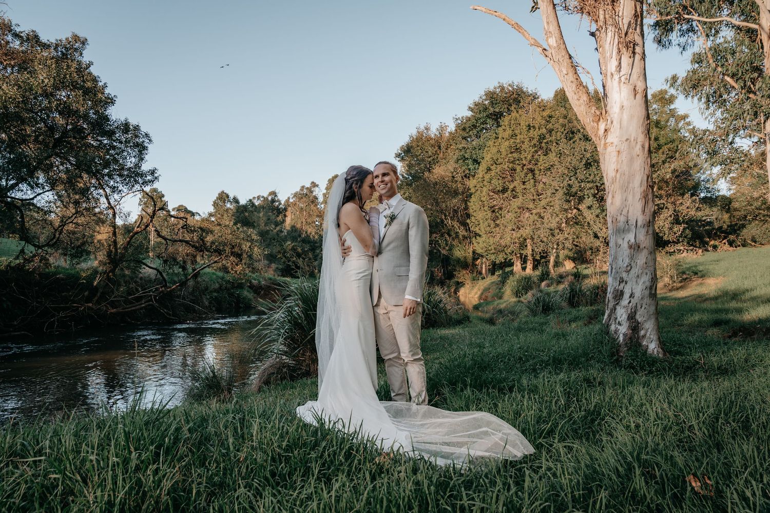 Wedding couple shares romantic moments in a golden sunset field by a lake during their outdoor countryside celebration.