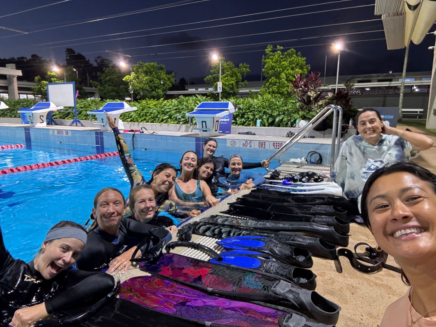 A group of scuba divers with gear gathered at night by an illuminated swimming pool with lane markers.