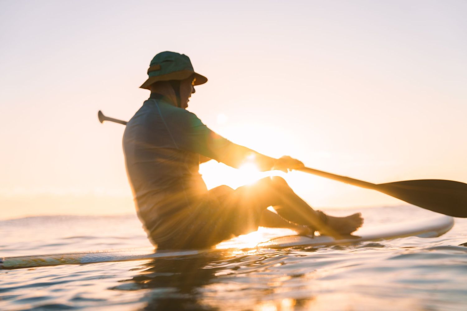 Silhouetted kayaker paddling on calm water at sunset with golden sunlight streaming through the frame.