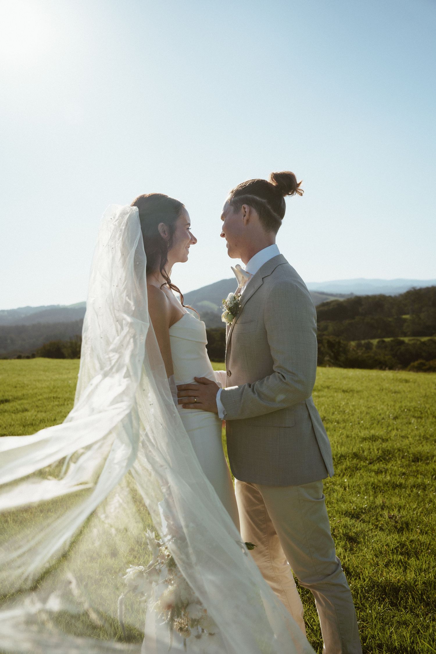 A bride and groom share an intimate moment on a hilltop meadow with flowing white veil and grey suit against mountain views.