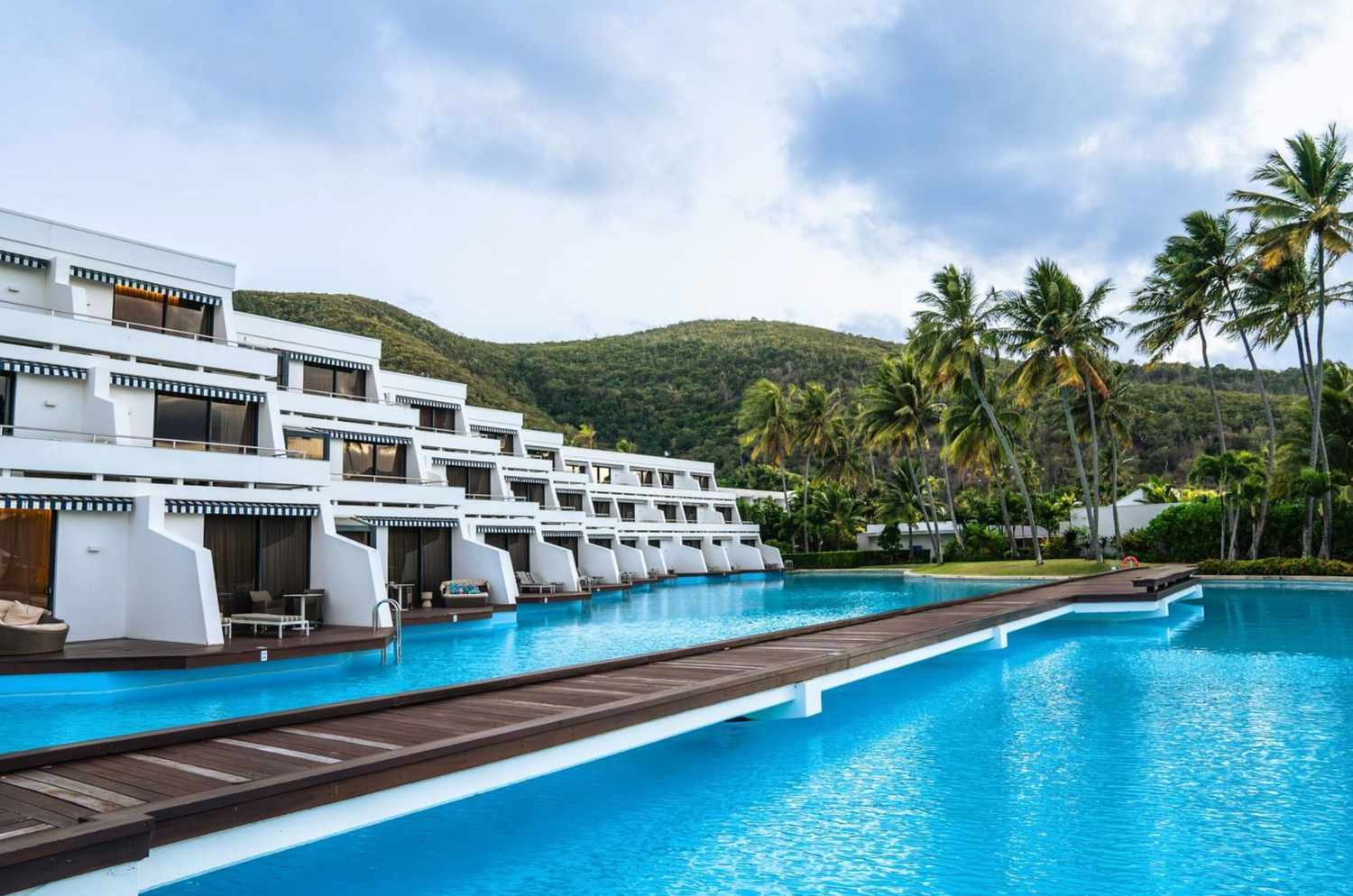 Luxury resort with modern white buildings and a long infinity pool overlooking a tropical palm-lined coastline.
