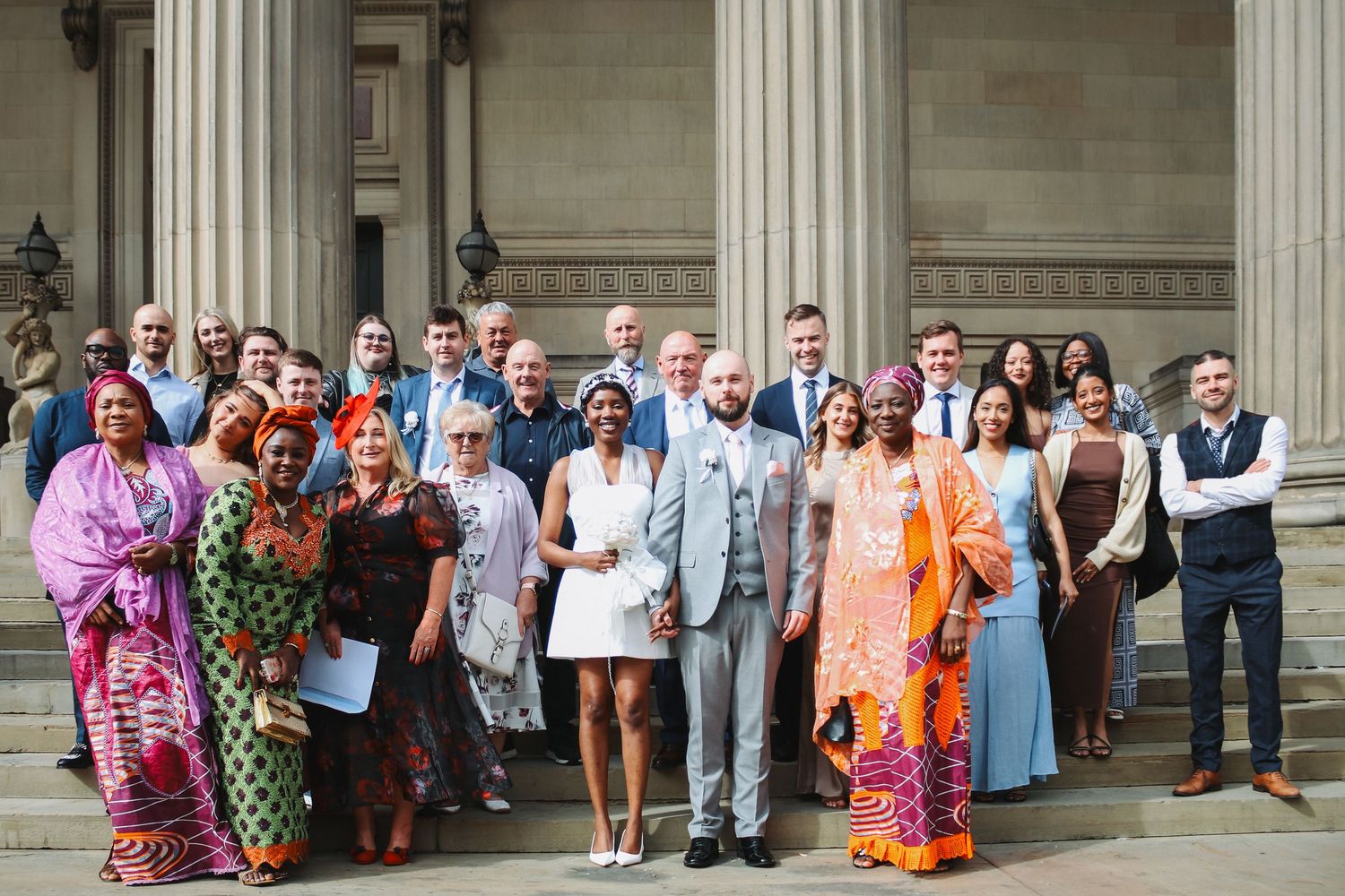Large wedding party group photo on stone steps with diverse guests in colorful attire.