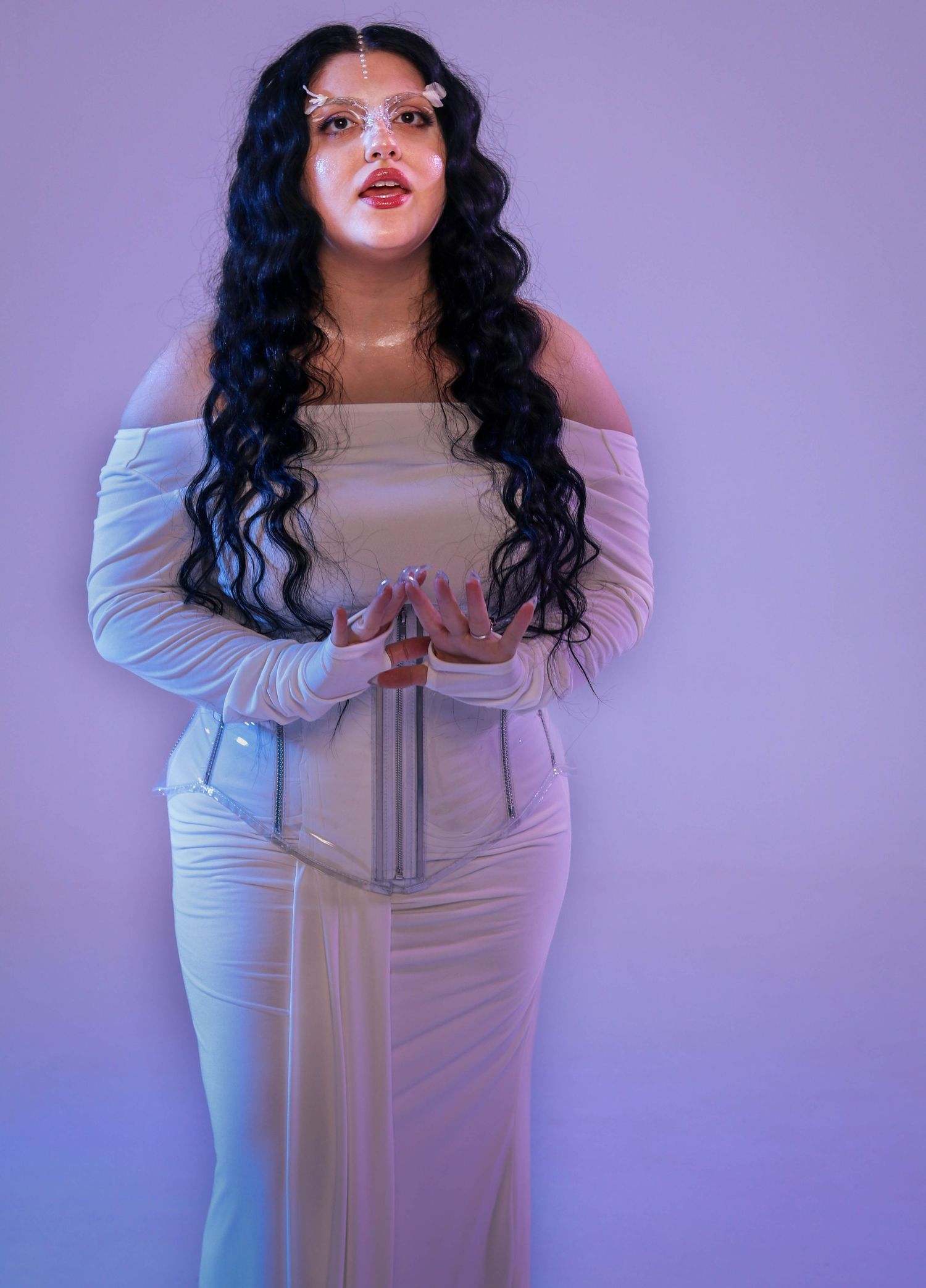 Woman with long black wavy hair posing in a white off-shoulder corset dress against a purple background.