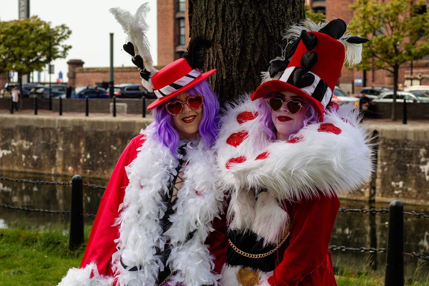 Two women in matching red and white costumes with feather boas and polka dot hats pose by a tree outdoors.