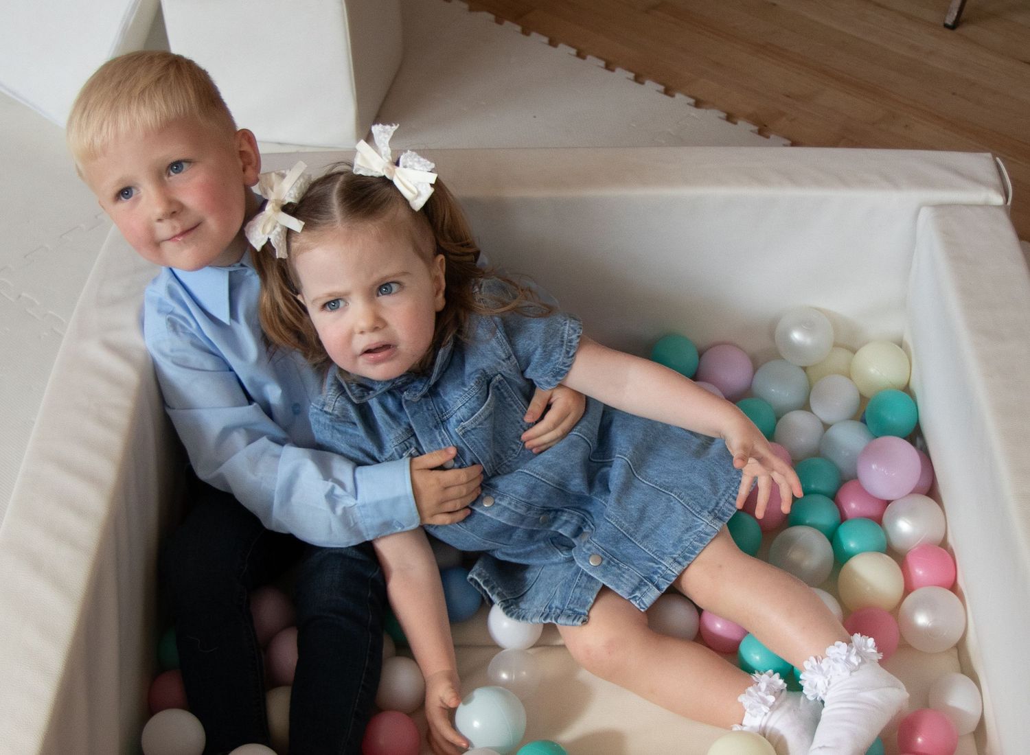 Two young children sitting together in a white ball pit filled with colorful pastel balls, both wearing blue outfits.