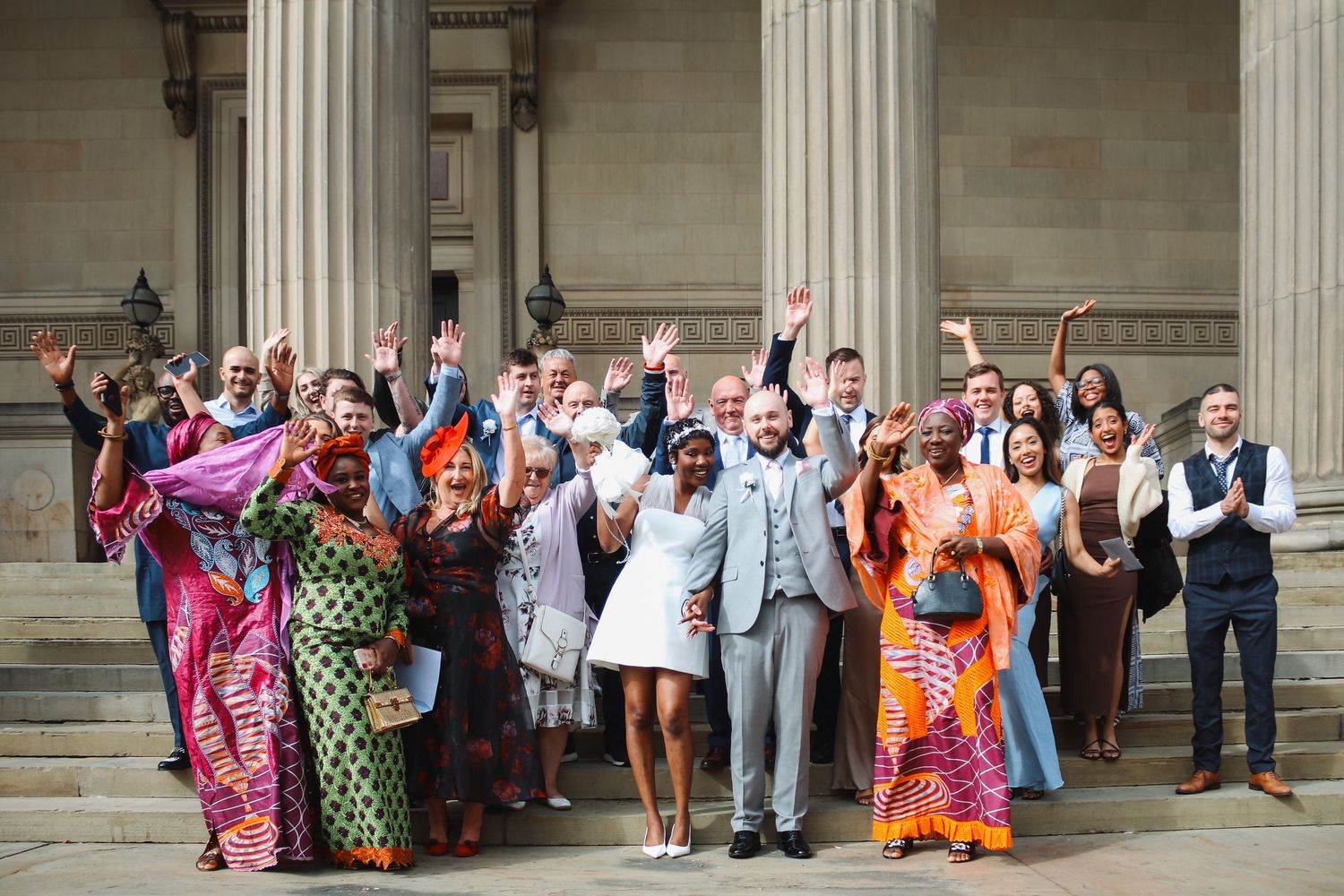 A joyful wedding party celebrates on steps of a grand building with colorful attire.