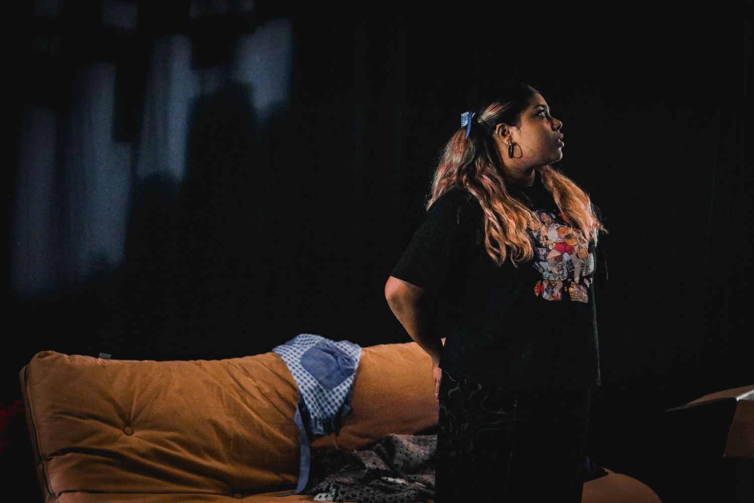 A young woman with long hair stands near a sofa in a dramatic dark stage performance setting.