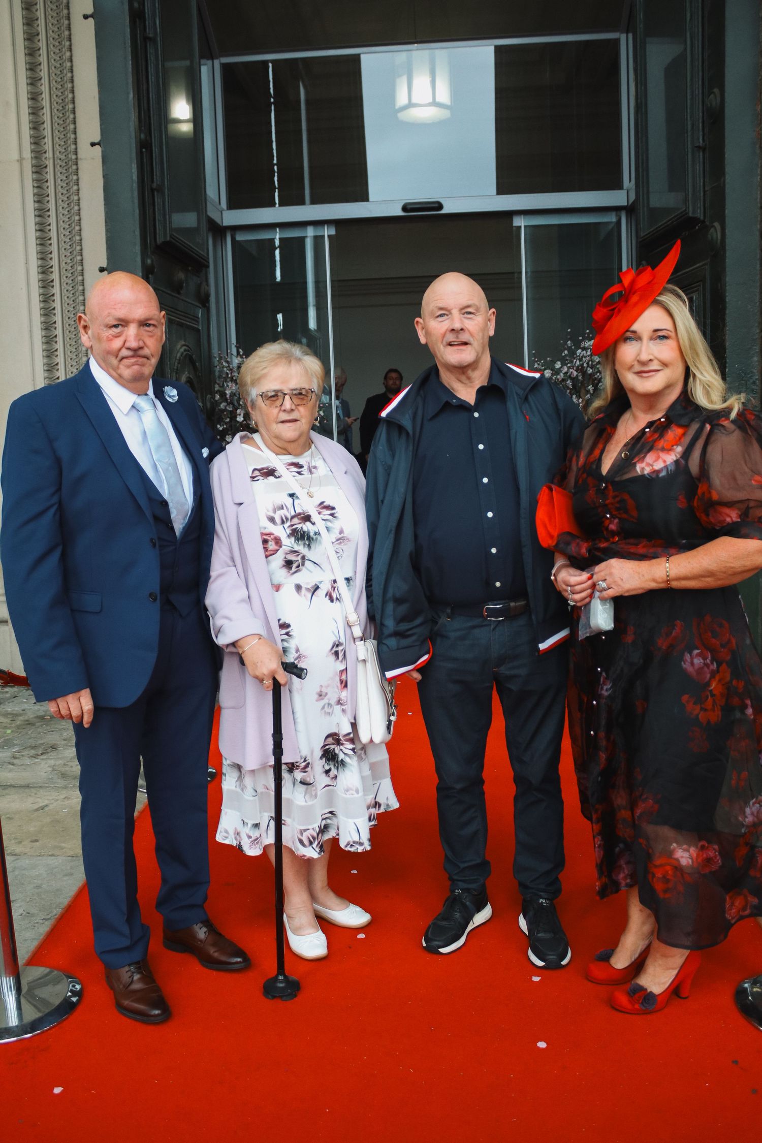 Four elegantly dressed adults pose on a red carpet outside a formal event venue.