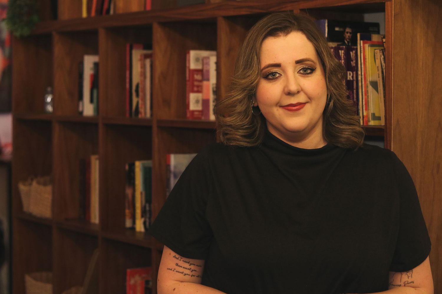 Woman in black t-shirt smiling in front of wooden bookshelves filled with colorful books in a cozy bookstore.