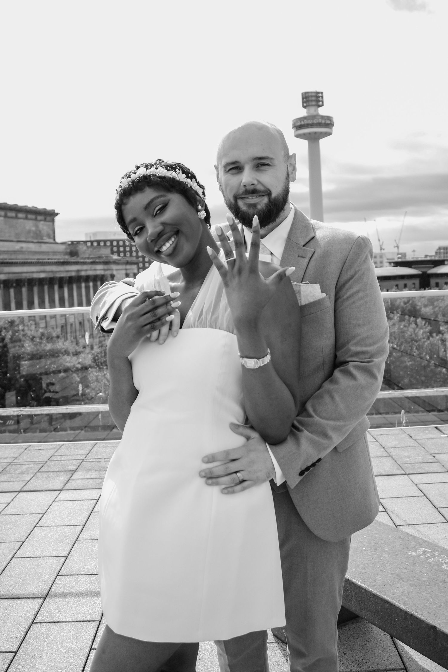 Black and white photo of newlyweds showing wedding rings outdoors with tower in background.