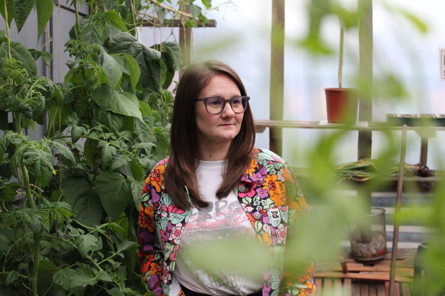 Young woman with glasses in colorful floral jacket surrounded by lush green plants in a greenhouse setting.