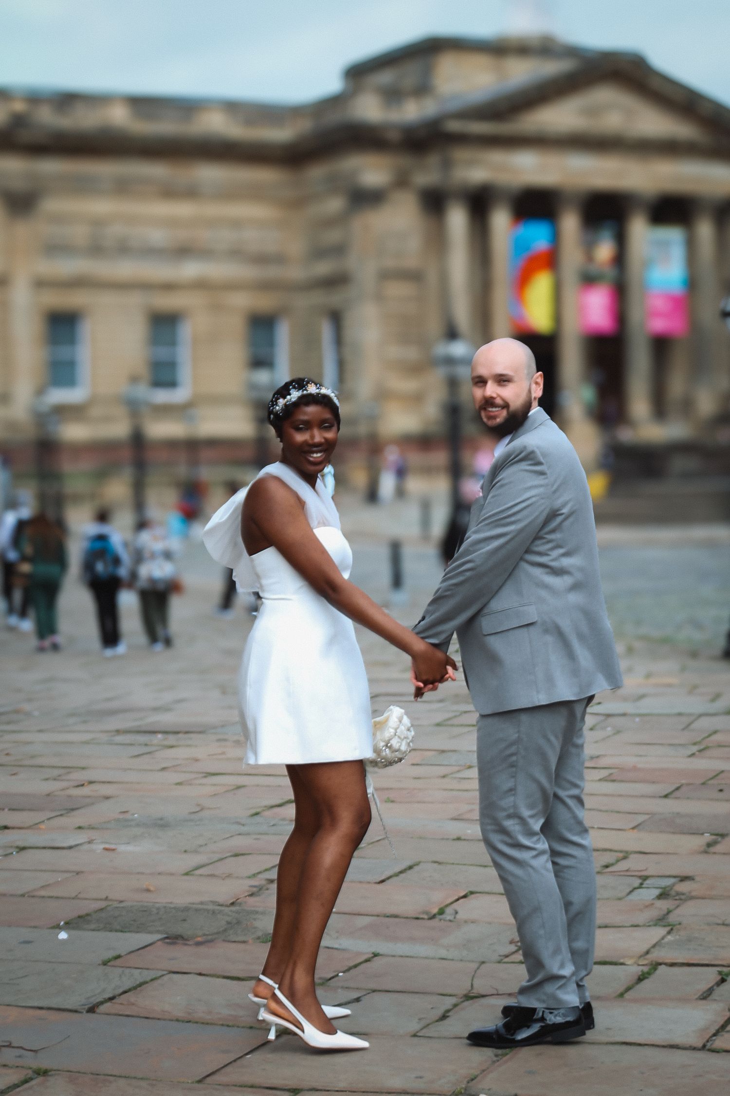 Interracial couple in wedding attire holding hands outdoors, bride in white mini dress, groom in gray suit, historic building backdrop.