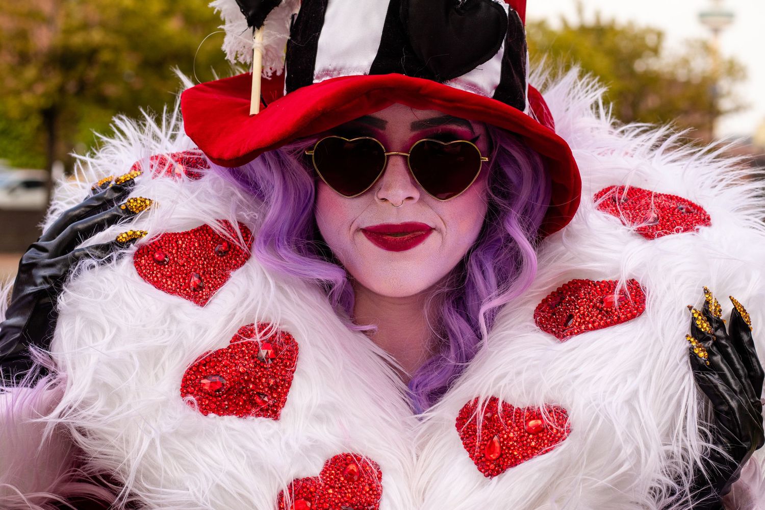Woman in Queen of Hearts costume with red hat, heart-covered fur coat, purple hair, and sunglasses at cosplay event.
