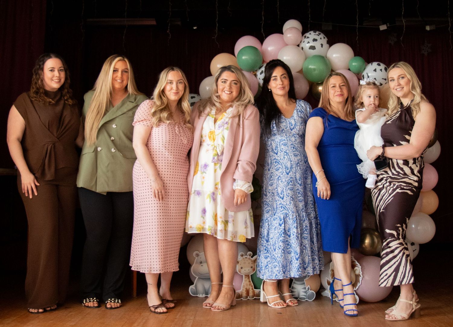 Group of seven women posing together at a baby shower with pastel balloon arch backdrop.