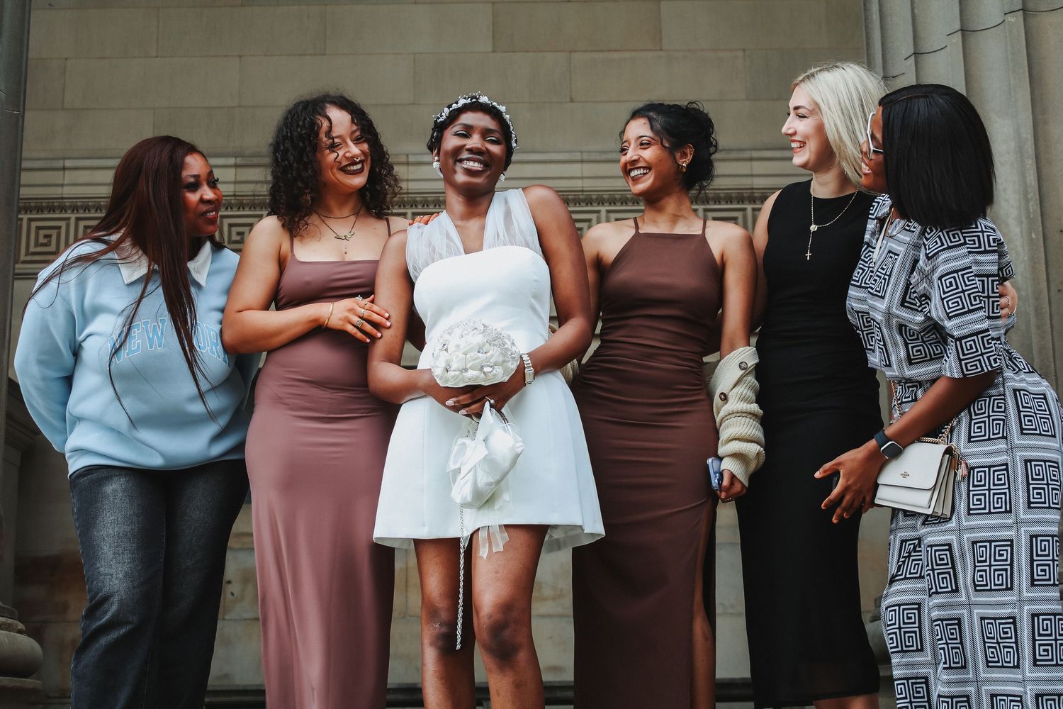 Bride in white mini dress laughs with bridesmaids in mauve gowns outside stone building.