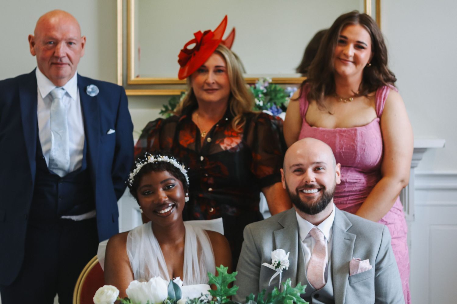 A joyful wedding group photo with bride and groom seated, flanked by three smiling guests in formal attire.