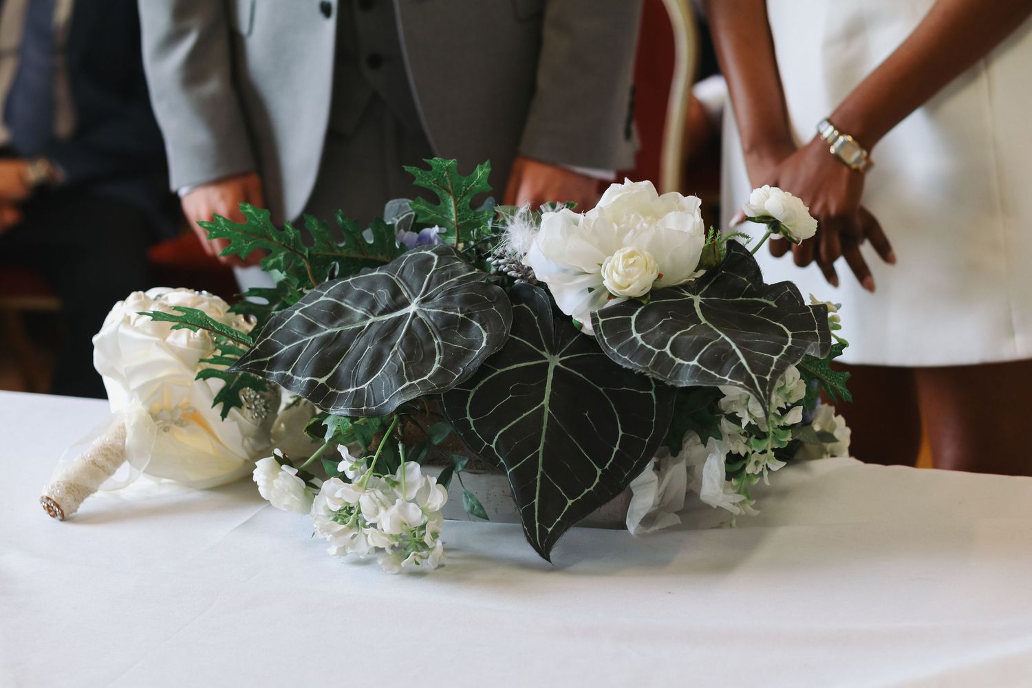 Elegant wedding centerpiece with white roses and dark tropical leaves in a silver bowl on a white tablecloth.