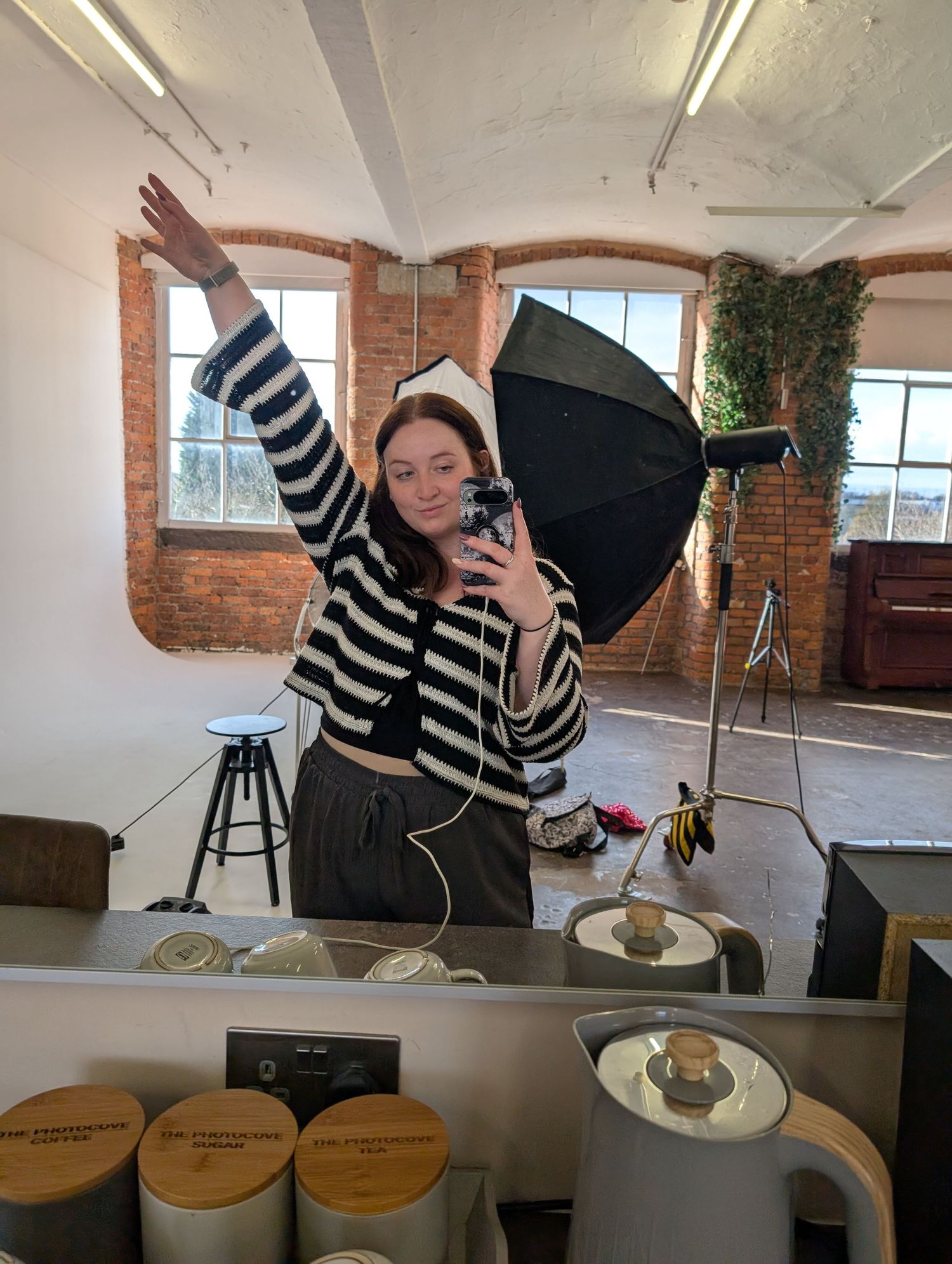 Woman in striped cardigan takes mirror selfie in photography studio with softbox light and brick arched windows.