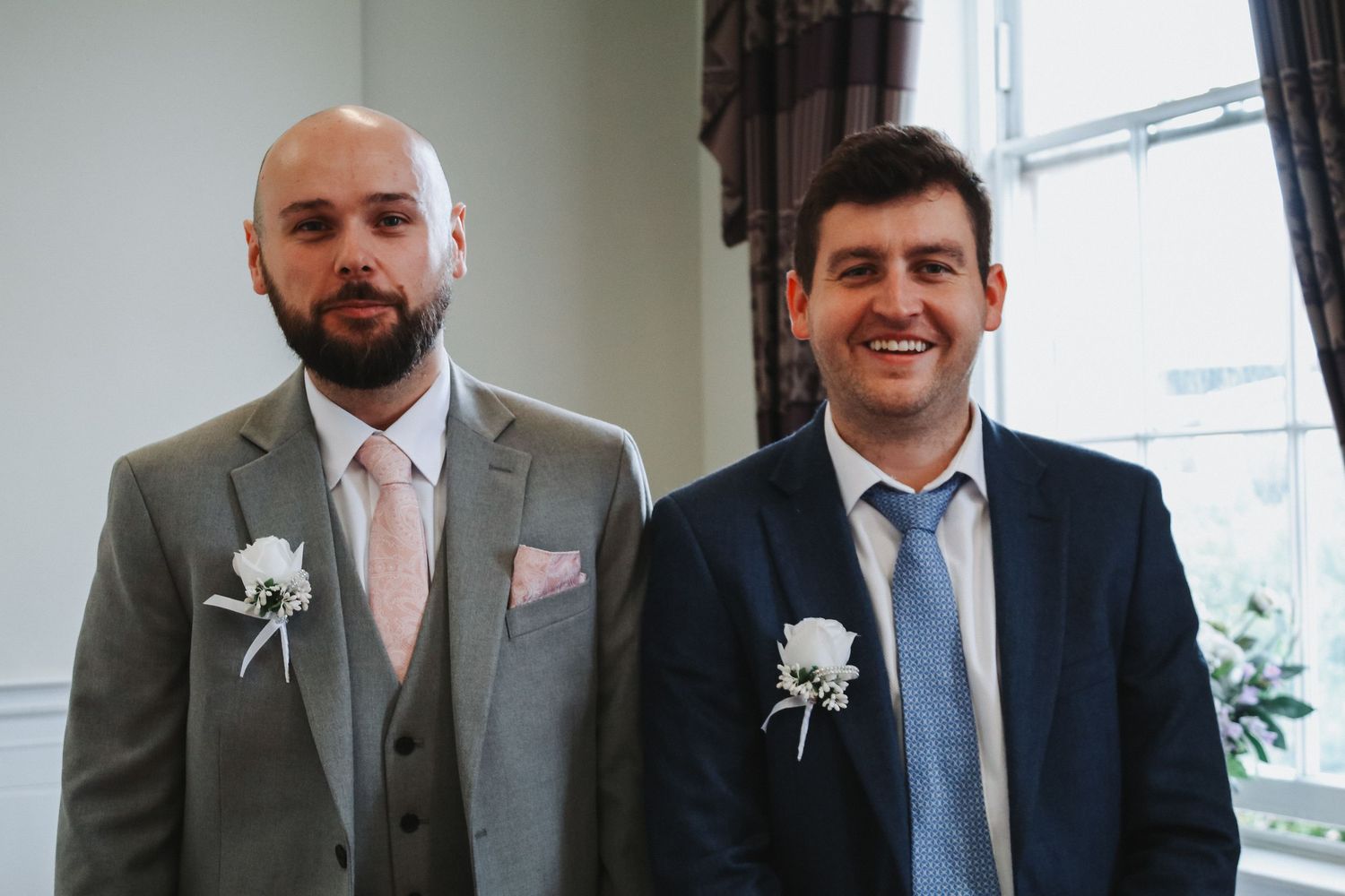 Two smartly dressed men in suits with white boutonnieres smiling at a wedding ceremony indoors.