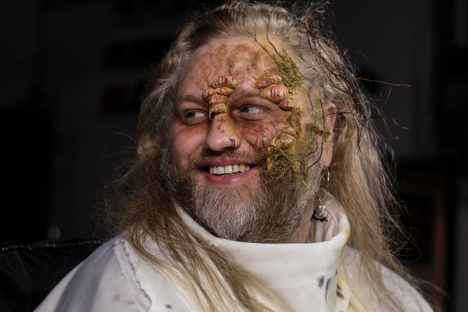 Man with long hair and beard wearing elaborate fantasy creature makeup and white costume, smiling at camera.