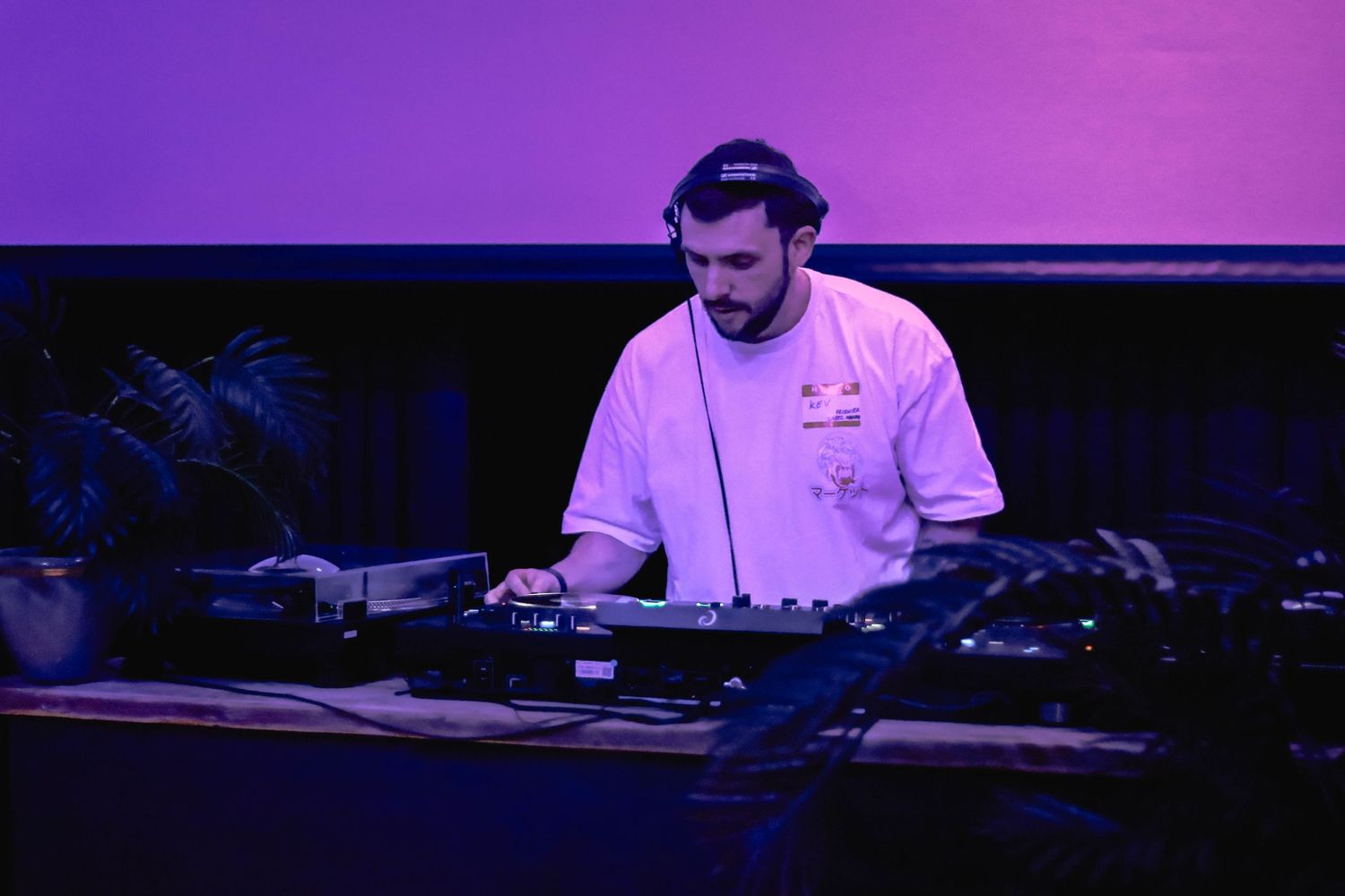 Male DJ wearing headphones and a white shirt performing at a DJ booth under purple and pink stage lighting.