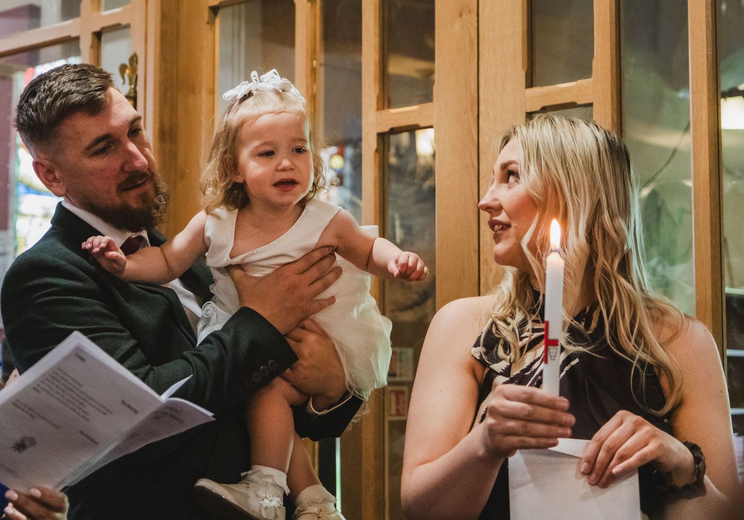 Bearded man in green suit holding baby girl beside blonde woman at social gathering.