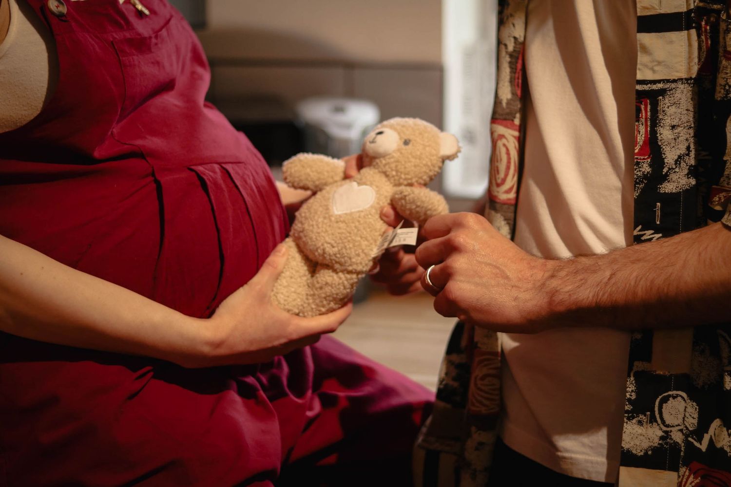 Pregnant woman in red dress holding a teddy bear with a man beside her, symbolizing parenthood and new baby anticipation.
