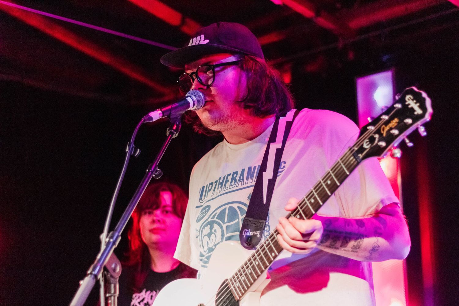 Tattooed guitarist singing into microphone on stage wearing white t-shirt and cap at live music venue.