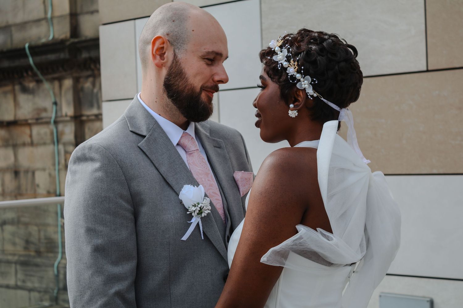 Interracial couple in wedding attire gazing lovingly at each other, bride in white gown with floral headpiece, groom in gray suit.